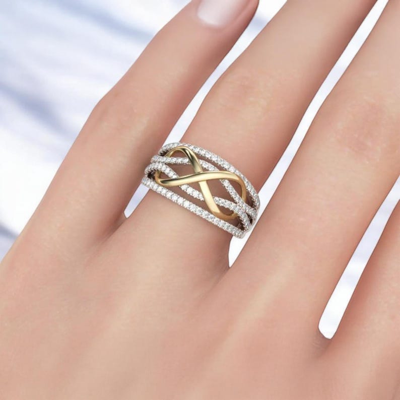 K&ouml;nnte beinhalten: Ein gold-silberner Ring mit einem Unendlichkeits-Symbol-Design. Der Ring hat ein goldenes Unendlichkeits-Symbol in der Mitte, umgeben von silbernen B&auml;ndern, die mit kleinen, klaren Edelsteinen besetzt sind. Der Ring befindet sich an einem Finger.
