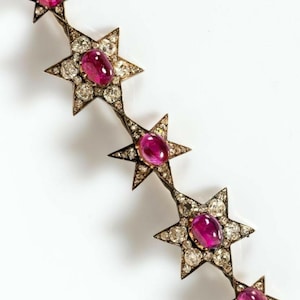 May include: A vintage starburst brooch featuring five star-shaped elements. Each star is adorned with a central oval ruby and surrounded by sparkling diamonds. The brooch has a dark metal setting and is set against a white background.