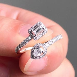 May include: Two silver rings with clear gemstones. One ring features a rectangular gemstone surrounded by smaller stones. The other ring has a pear-shaped gemstone, also surrounded by smaller stones. The rings are on a finger.