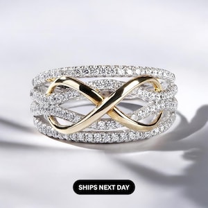 K&ouml;nnte beinhalten: Ein silberfarbener Ring mit einem goldfarbenen Unendlichkeitszeichen in der Mitte. Der Ring ist mit kleinen, klaren Steinen besetzt. Die Worte "SHIPS NEXT DAY" werden in einem schwarzen Rechteck unten angezeigt.