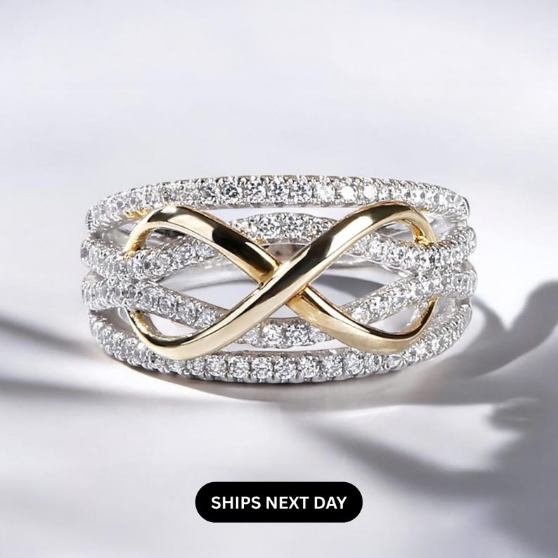 K&ouml;nnte beinhalten: Ein silberfarbener Ring mit einem goldfarbenen Unendlichkeitszeichen in der Mitte. Der Ring ist mit kleinen, klaren Steinen besetzt. Die Worte "SHIPS NEXT DAY" werden in einem schwarzen Rechteck unten angezeigt.