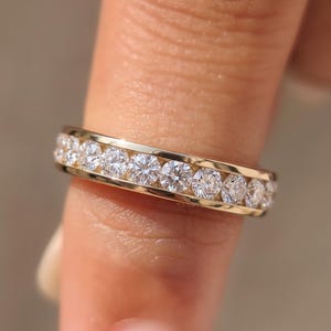 May include: A gold band ring featuring a channel setting with a continuous row of round, clear, faceted stones. The metal has a polished finish, and the stones are arranged side-by-side along the center of the band, creating a uniform, sparkling appearance.
