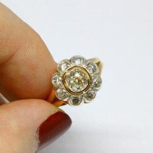 May include: A gold ring featuring a central diamond surrounded by a halo of smaller diamonds, creating a flower-like design. The ring is held against a white background, showcasing its intricate details and sparkle.