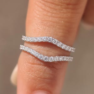 May include: A double-band ring set crafted from silver-toned metal, featuring two curved bands each encrusted with a row of small, clear, round-cut stones that catch the light, creating a shimmering, continuous pavé effect across the surface.