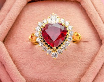 Könnte beinhalten: Ein goldener Ring mit einem großen herzförmigen roten Edelstein in der Mitte, umgeben von einem Kranz aus kleinen klaren runden Steinen und einem einzelnen klaren Stein im Baguetteschliff an der Oberseite, vor einem strukturierten rosa Hintergrund.