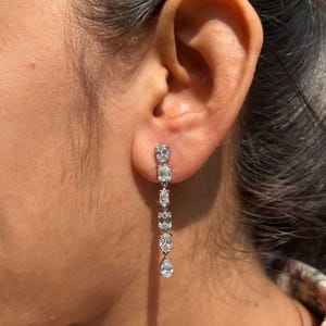 May include: A pair of long, silver dangle earrings with multiple clear, oval-shaped stones. The earrings are shown on a person's ear.