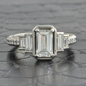 May include: A platinum engagement ring with an emerald-cut center diamond, flanked by two smaller baguette diamonds. The band features a pave setting of small diamonds. The ring is set against a textured, dark background.