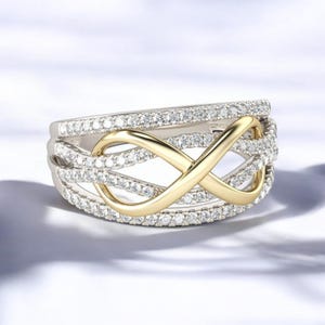 K&ouml;nnte beinhalten: Ein silberfarbener Ring mit einem goldfarbenen Unendlichkeitssymbol in der Mitte. Der Ring ist mit kleinen, klaren Edelsteinen besetzt. Das Unendlichkeitssymbol steht im Mittelpunkt, vor dem Hintergrund der funkelnden Edelsteine.