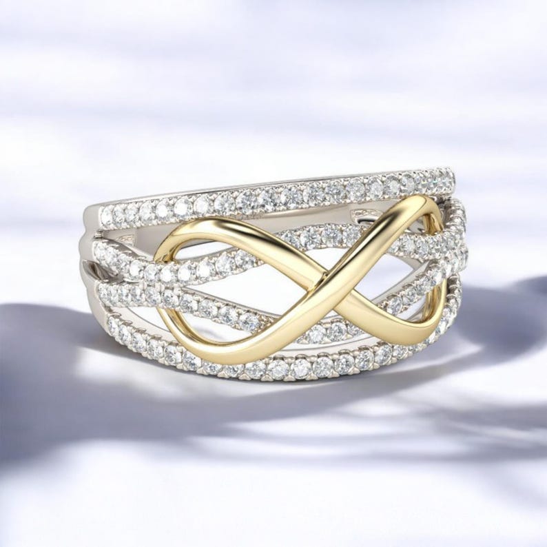 K&ouml;nnte beinhalten: Ein silberfarbener Ring mit einem goldfarbenen Unendlichkeitssymbol in der Mitte. Der Ring ist mit kleinen, klaren Edelsteinen besetzt. Das Unendlichkeitssymbol steht im Mittelpunkt, vor dem Hintergrund der funkelnden Edelsteine.