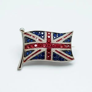 May include: A silver-toned brooch in the shape of the Union Jack flag. The flag design features red and blue gemstones set in a silver frame. The brooch has a pin on the back for secure fastening.