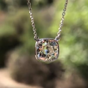 May include: A silver necklace with a large, square-cut diamond pendant. The diamond is set in a bezel setting and sparkles with multiple facets. The chain is delicate and hangs against a blurred green background.