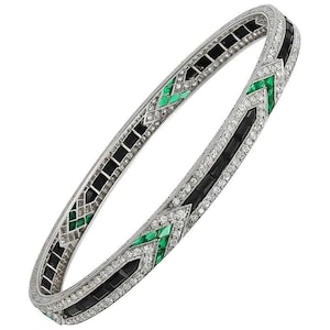 Puede incluir: Un brazalete de plata con un diseño geométrico con piedras preciosas negras y verdes. El brazalete está engastado con diamantes y esmeraldas.
