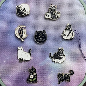 May include: Nine enamel pins featuring black cats in various poses. The pins are on a purple and white fabric background. The pins are all different designs, including a cat with a crescent moon, a cat with a skull, and a cat with flowers.