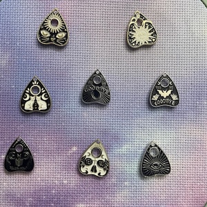May include: Eight black and white enamel pins shaped like a witch's  'planchette' or 'ouija board' pointer. The pins feature various designs including a sun, a moon, a cat, a bat, and the words 'Yes', 'No', and 'Goodbye'.