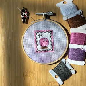 May include: A cross-stitch embroidery hoop featuring a pink and white design of a crystal ball. A coffin-shaped needle minder and spools of embroidery floss in various colors, including gray, brown, purple, and light blue, are arranged around the hoop on a wooden surface.