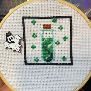 May include: A cross-stitch embroidery of a green potion bottle with a cork stopper, surrounded by green four-leaf clover designs, framed in black.