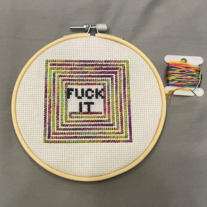 Puede incluir: Un bordado de punto de cruz en un aro de madera presenta el texto "FUCK IT" en negro. El diseño está rodeado por un patrón cuadrado colorido. Una bobina de hilo multicolor se encuentra junto al aro.