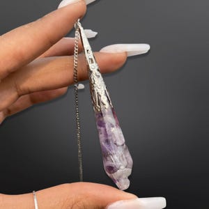 May include: A pointed, cone-shaped pendulum pendant featuring a collection of small, tumbled purple amethyst and clear quartz stones encased in a silver-toned metal cap with intricate cut-out scrollwork, suspended from a thin silver-toned chain.