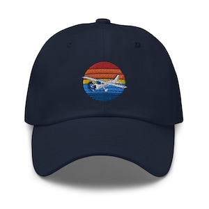 May include: A navy blue baseball cap with an embroidered design of a small airplane flying in front of a sunset. The sunset is depicted in shades of orange, yellow, and blue.