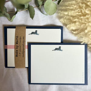 May include: A set of notecards with cream-coloured paper and navy blue borders. Each card features a small illustration of a dog. The set is bound with a brown paper band that reads "Made by Marti".