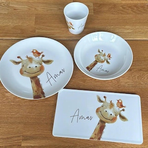 Könnte beinhalten: Ein weißes Keramik-Geschirrset mit einem Giraffen- und Vogeldesign. Das Set beinhaltet einen Teller, eine Schüssel, ein rechteckiges Tablett und eine Tasse. Der Name "Amar" ist auf jedem Stück geschrieben. Die Giraffe hat ein lächelndes Gesicht.