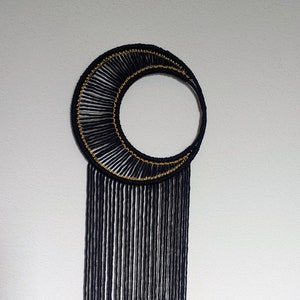 May include: Black and gold macrame wall hanging in the shape of a crescent moon with long fringe.