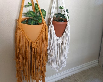 May include: Two macrame plant hangers with wooden rings, one is mustard yellow and the other is white. Both hangers have a plant in a terracotta pot.