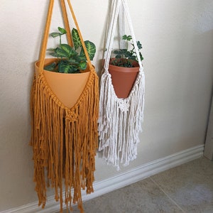 May include: Two macrame plant hangers with wooden rings, one is mustard yellow and the other is white. Both hangers have a plant in a terracotta pot.