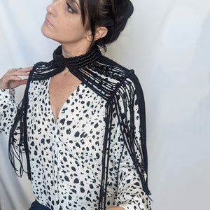 May include: A black fringe collar necklace with a long, flowing design. The necklace is made of a thick, woven cord and has a variety of fringe lengths. The necklace is worn over a white blouse with a black and white polka dot pattern.