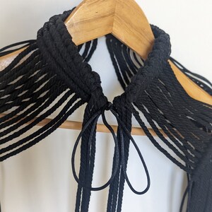May include: A black macrame choker necklace with long fringe tassels. The necklace is hanging on a wooden hanger.