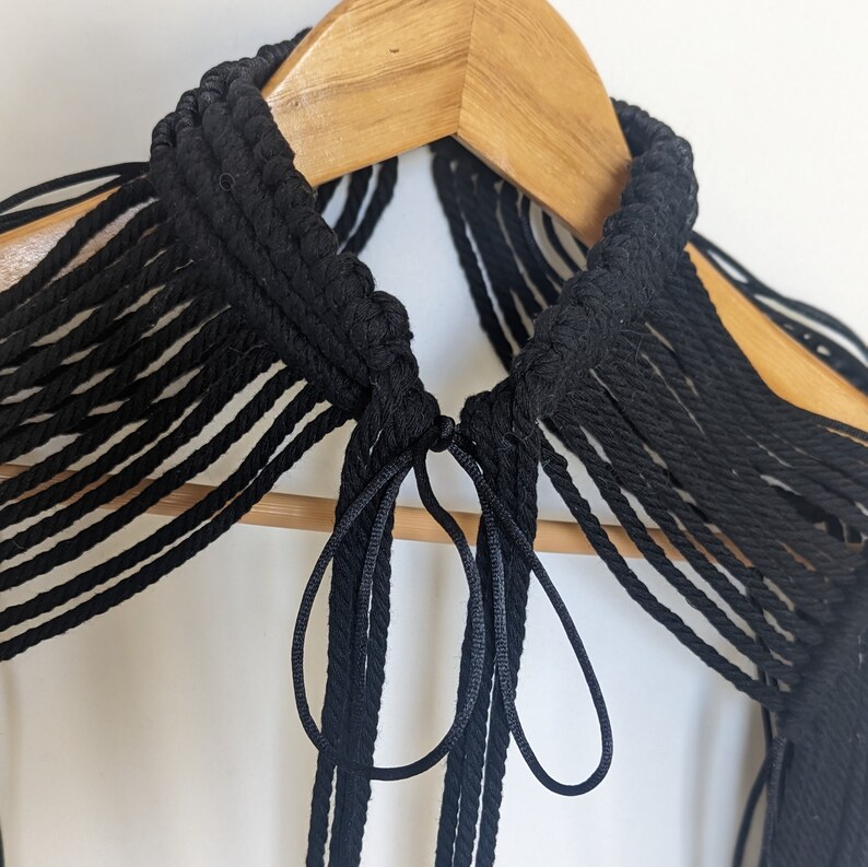 May include: A black macrame choker necklace with long fringe tassels. The necklace is hanging on a wooden hanger.
