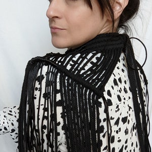 May include: A black macrame fringe scarf with a geometric pattern. The scarf is worn over a white and black animal print blouse.