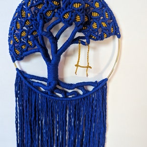 May include: A blue and gold macrame wall hanging in the shape of a tree with a swing hanging from a branch. The tree has a fringe of blue yarn hanging from the bottom.