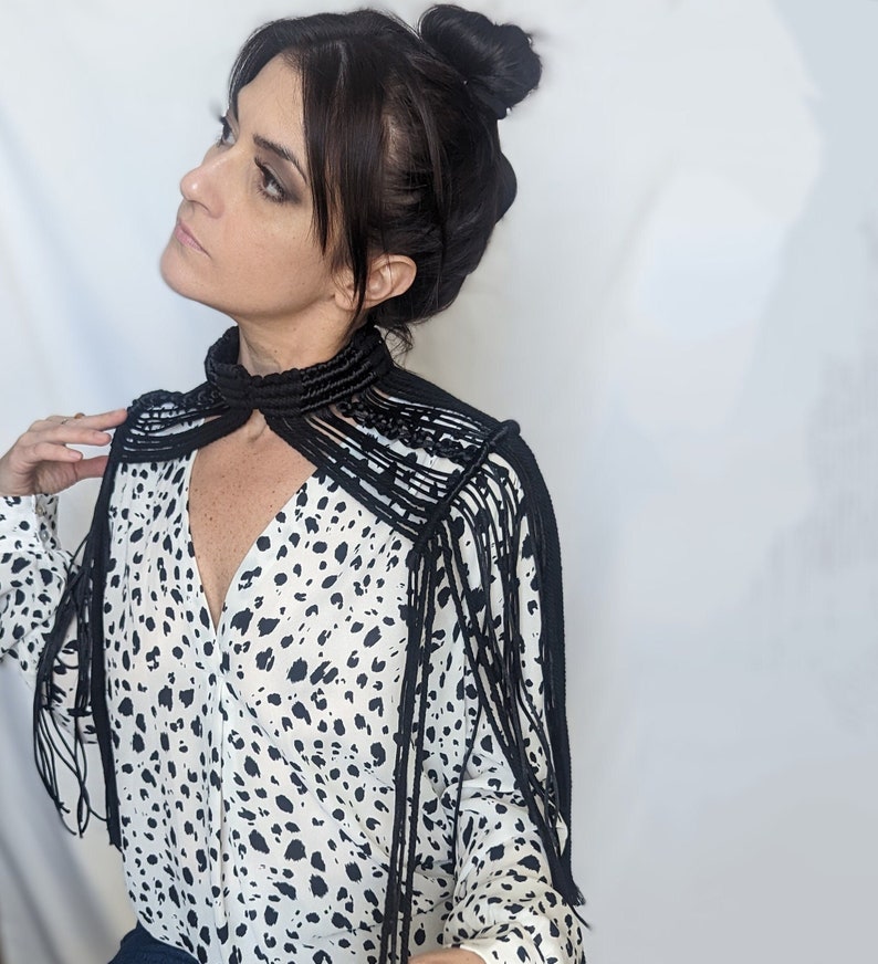 May include: A black fringe collar necklace with a long, flowing design. The necklace is made of a thick, woven cord and has a variety of fringe lengths. The necklace is worn over a white blouse with a black and white polka dot pattern.