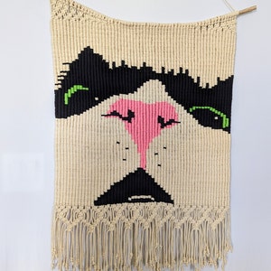 May include: A wall hanging made of macrame with a cat face design. The cat has black eyes with green accents, a pink nose, and a black mouth.