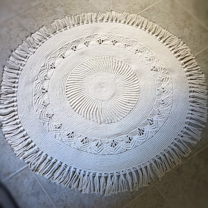 May include: A white round macrame rug with a fringe border. The rug has a circular pattern in the center and a geometric design around the edge.