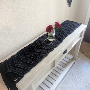 May include: Black macrame table runner with fringe on a light wood table with a vase of red roses.