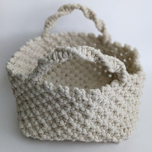 May include: A cream-colored crocheted basket with two handles. The basket is made of thick rope and has a woven pattern.