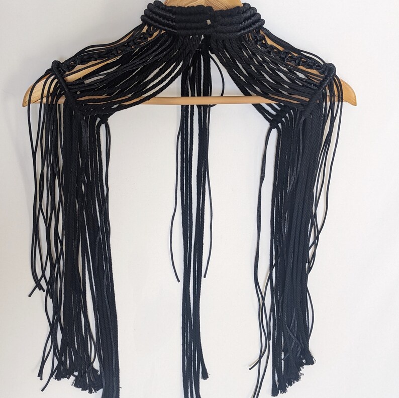 May include: Black macrame choker necklace with long fringe tassels. The necklace is made of multiple strands of cord that are knotted together to create a delicate and intricate design.