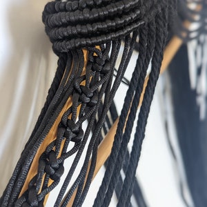 May include: Close-up of a black macrame wall hanging with a wooden dowel rod. The macrame is made with thick black cords and has a geometric pattern.