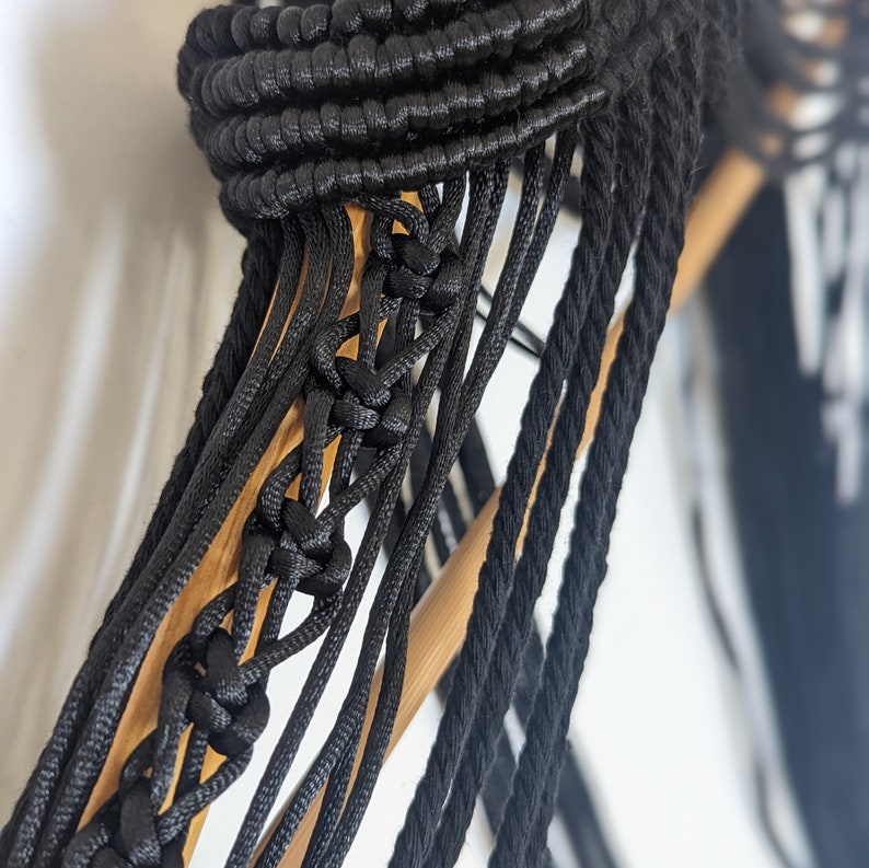 May include: Close-up of a black macrame wall hanging with a wooden dowel rod. The macrame is made with thick black cords and has a geometric pattern.