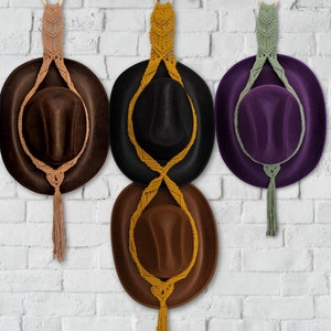 May include: Three brown, black, and purple hats with macrame cord hanging from them. The hats are hanging on a white brick wall.
