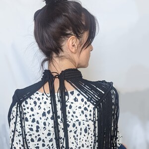 May include: A black rope necklace with long fringe strands extending from the shoulders. The necklace is worn over a white and black animal print blouse.