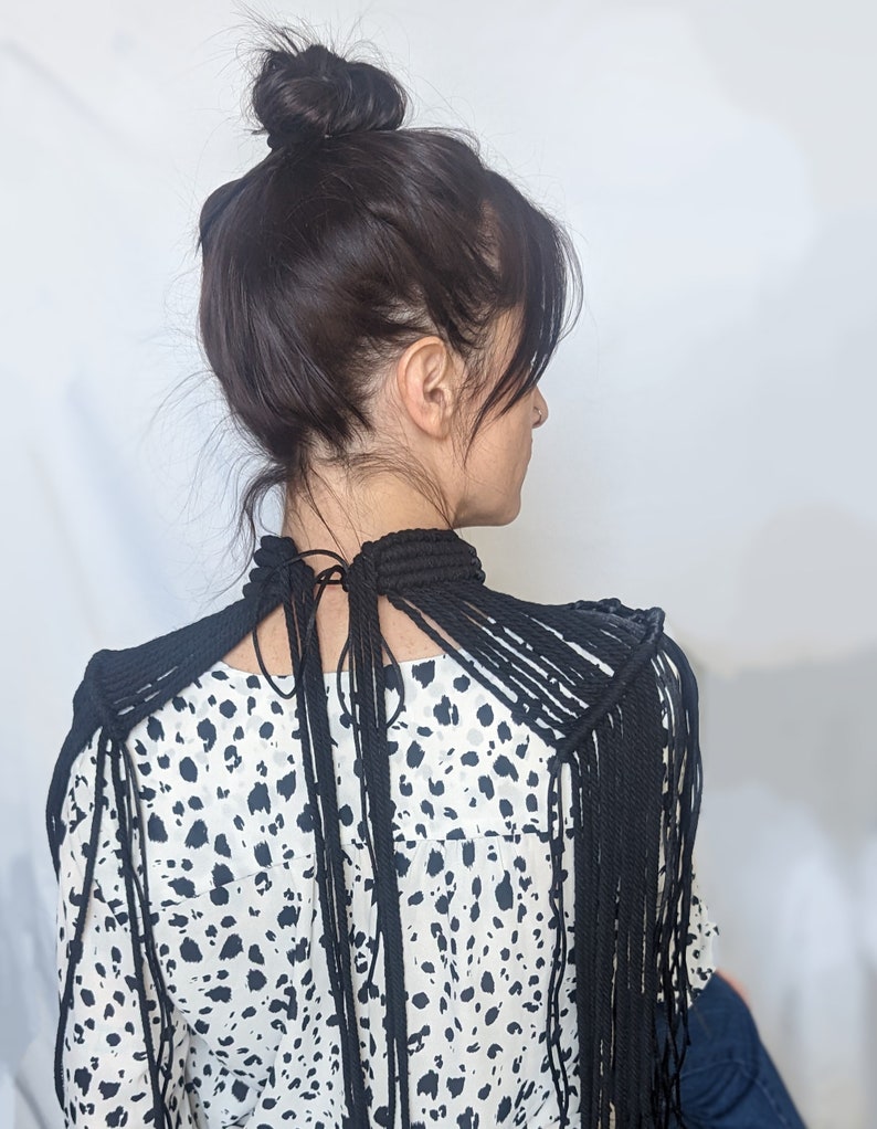 May include: A black rope necklace with long fringe strands extending from the shoulders. The necklace is worn over a white and black animal print blouse.