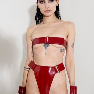 Puede incluir: Un conjunto de lencer&iacute;a de charol rojo con un top bandeau, bragas de cintura alta y pu&ntilde;os a juego. El conjunto presenta hebillas y herrajes plateados.