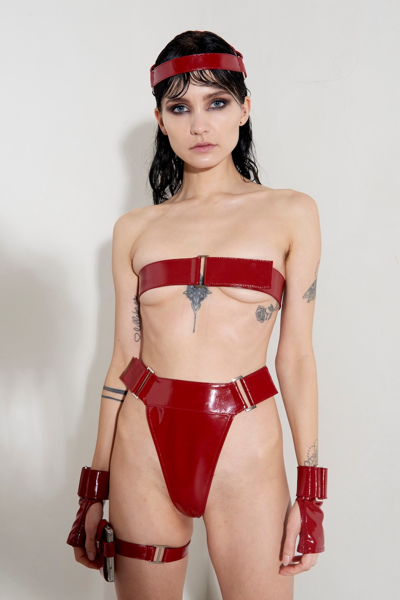 Puede incluir: Un conjunto de lencer&iacute;a de charol rojo con un top bandeau, bragas de cintura alta y pu&ntilde;os a juego. El conjunto presenta hebillas y herrajes plateados.