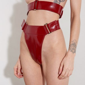 Puede incluir: Ropa interior de talle alto de cuero rojo patentado con una hebilla dorada y una pierna de corte alto.
