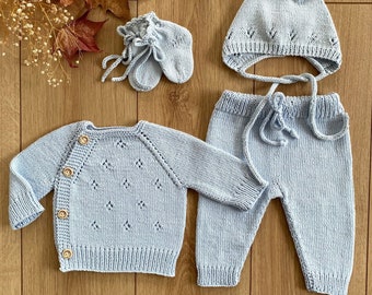 Op de afbeelding: Een lichtblauwe gebreide baby-outfitset met een trui, broek, muts en slofjes. De trui heeft een knoopsluiting en een delicaat patroon. De broek heeft een trekkoord. De muts heeft een eenvoudig ontwerp met een strik.