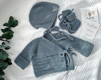 Op de afbeelding: Een set babykleding in lichtblauw, inclusief een trui met capuchon, een wikkeltrui en slofjes. De kleding is gemaakt van zachte, gebreide stof en perfect voor een pasgeboren baby.