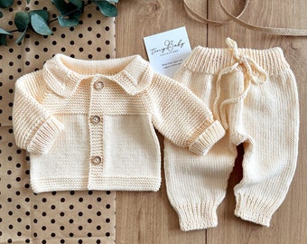Op de afbeelding: Een crèmekleurige gebreide baby-outfit met een vest en broek. Het vest heeft een knoopsluiting en een Peter Pan-kraag. De broek heeft een trekkoord.